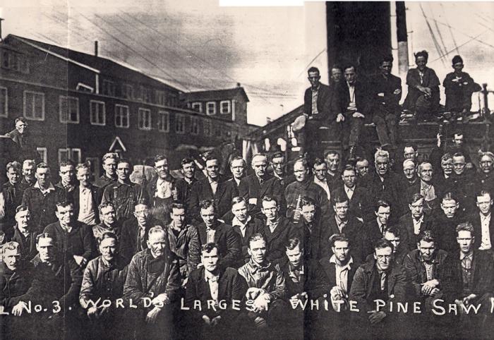 Group of workers standing in from of lumber yard in 1925. Solemn.