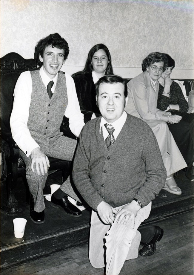 Man on bended knee faces the camera, well dressed wearing a tie under a cardigan. Behind him on a bench sits a well dressed Charlie Heymann with several people along side him on the bench. 