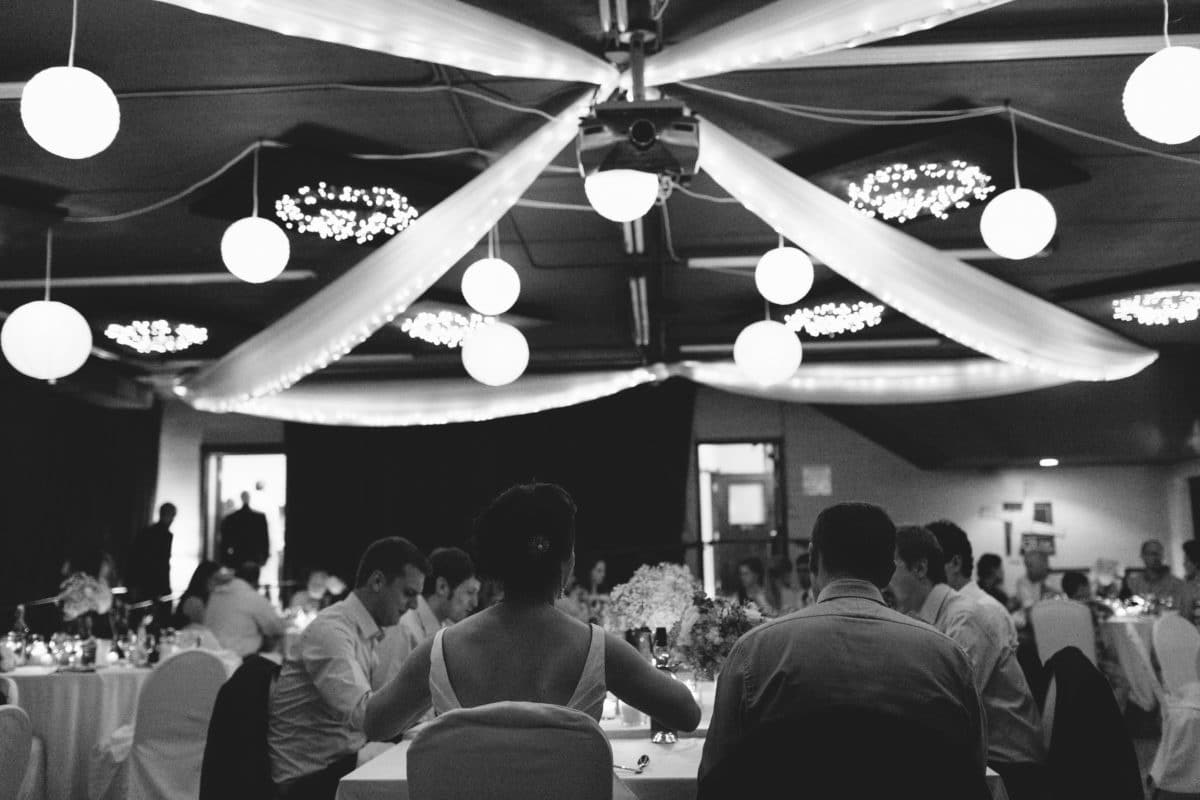 Black and white photo of a large hall with cheery swathes of white fabric draped from the ceiling, white paper lanterns hang cheerily. A crowd of fancily dressed folks eat at tables covered in white.