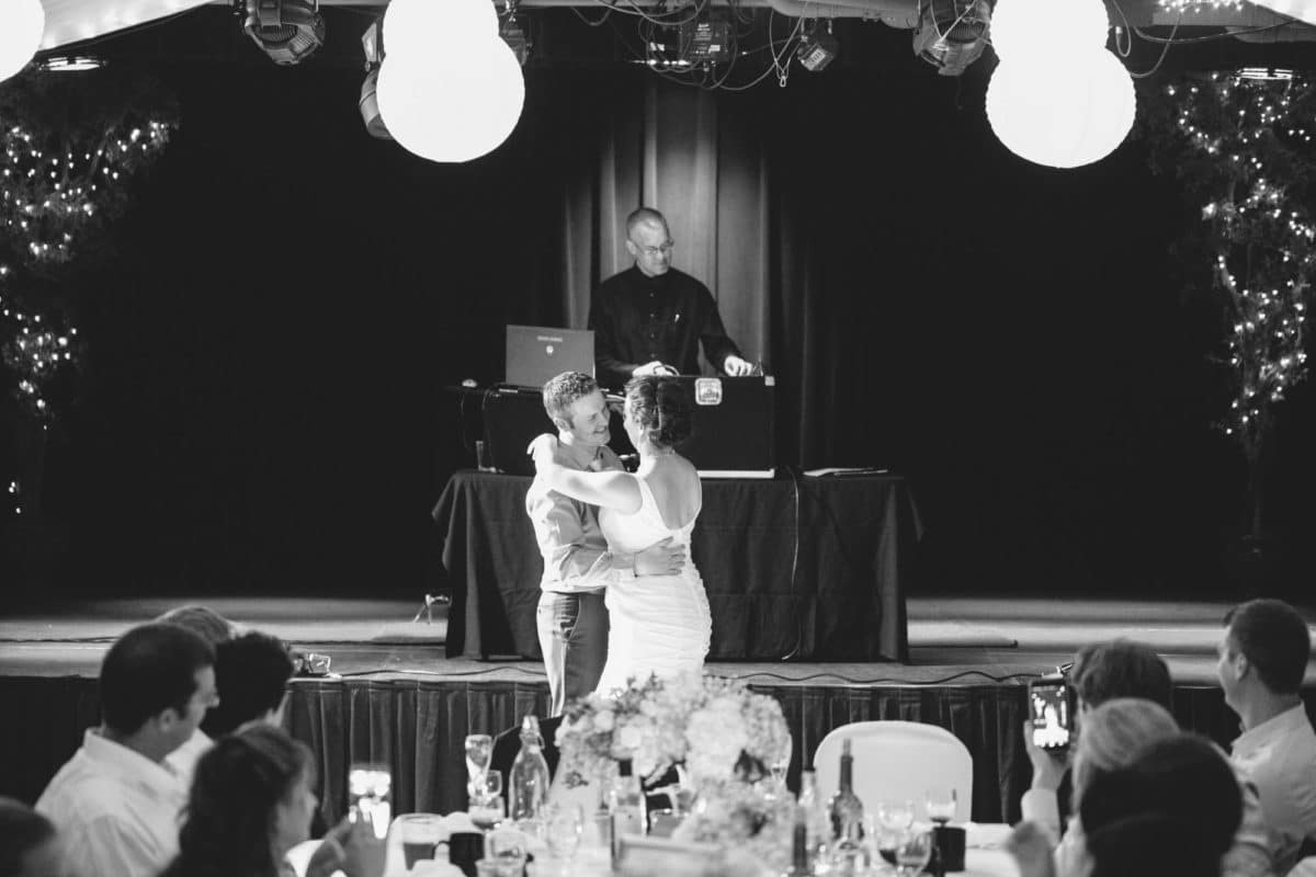 Wedding couple are having their first dance as a married couple in a lovely decorated hall. In the background a DJ stands on a stage, in the foreground, a white table is laden with crystal, goblets, wine bottles, and flowers. The couple only have eyes for each other. 